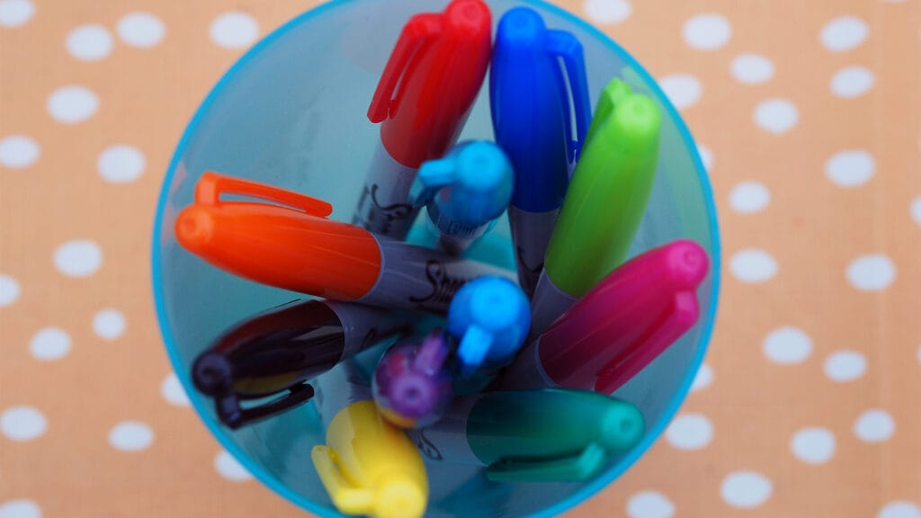 Erivärisiä tusseja sinisessä mukissa pilkullisen pöytäliinan päällä.